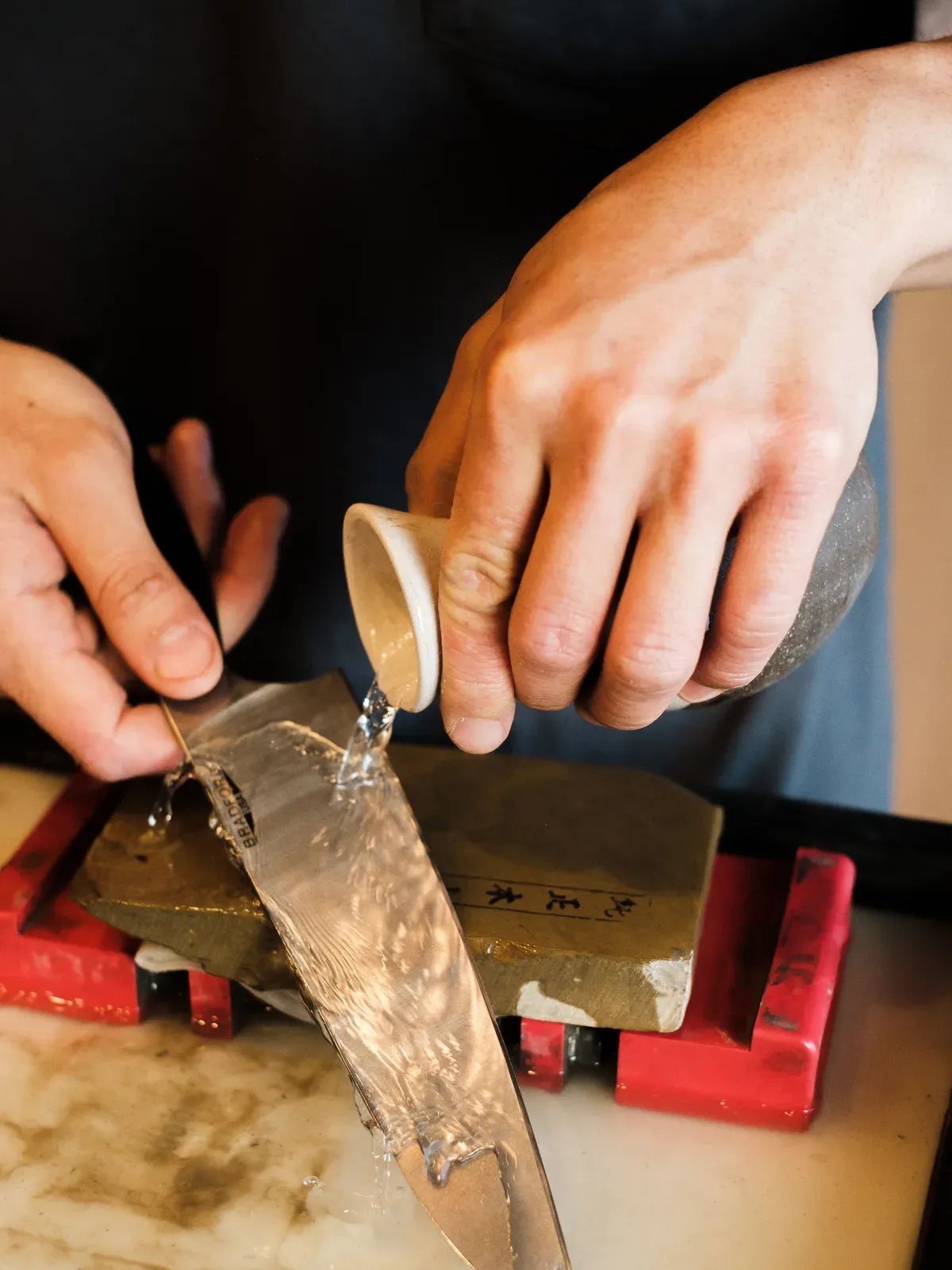 Two hands honing a blade on a wet whetstone, water running over the stone