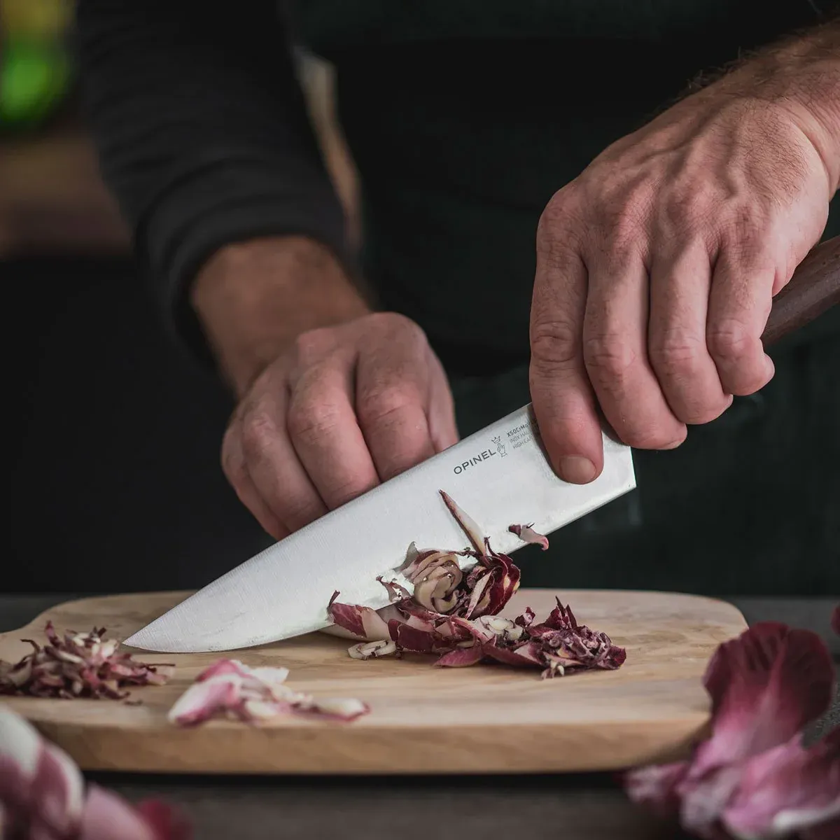A maker drawing a chef’s knife through radicchio on a worn cutting board, full attention on the cut